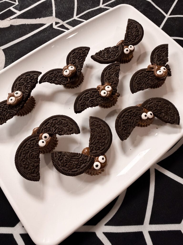 A white tray holds several bat-shaped cookies made from chocolate sandwich cookies and candy eyes, arranged on a black spiderweb-patterned tablecloth—a delightfully spooky treat that’s perfect for your list of 10 Easy Halloween Ideas.