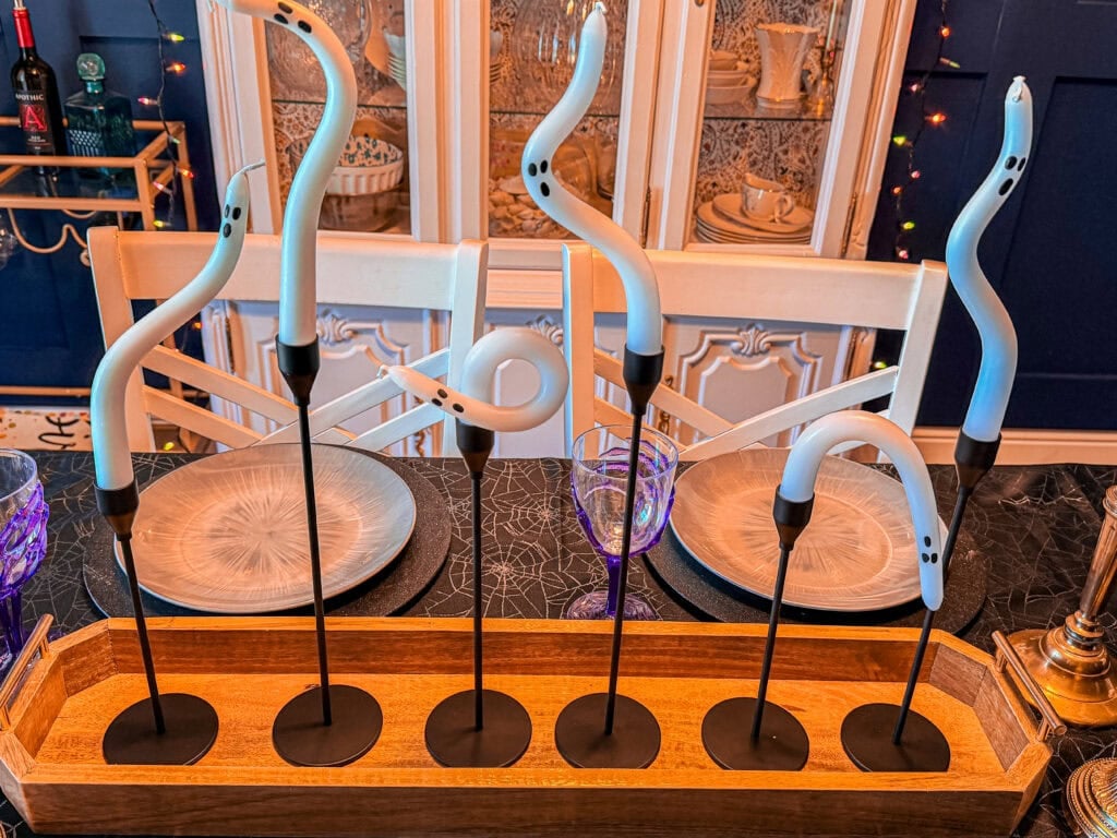 Six white, snake-like sculptures with black bases are displayed on a wooden tray, sitting on a dining table set with gray plates and purple glasses—one of 10 Easy Halloween Ideas to instantly add a spooky touch. A cabinet with dishes and string lights is in the background.