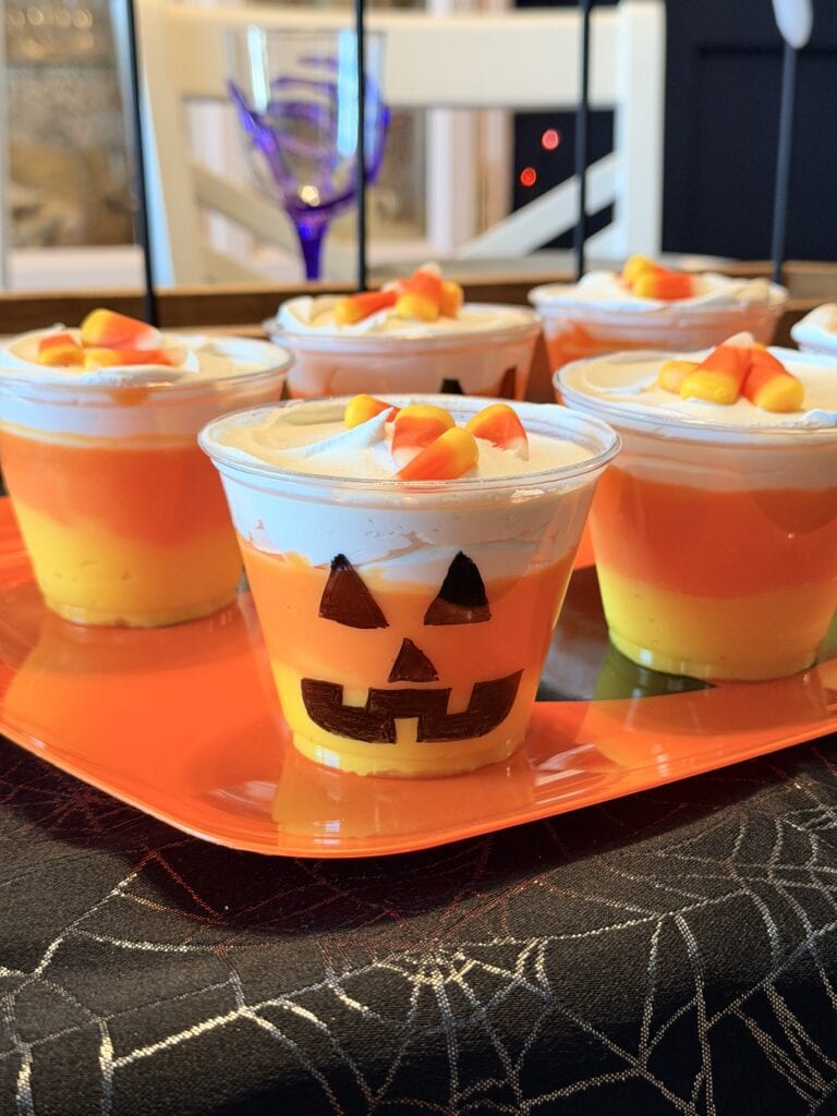 Plastic cups filled with layered yellow, orange, and white dessert, topped with candy corn, offer a festive touch to your party spread. One cup even features a jack-o-lantern face for added fun. Set on an orange tray over a black, spiderweb-patterned tablecloth, this treat is perfect if you’re searching for 10 Easy Halloween Ideas to impress your guests.