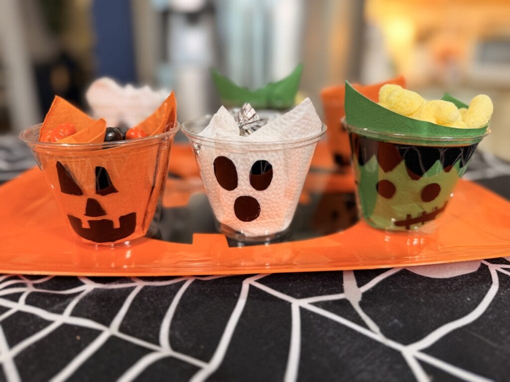 Three clear cups decorated for Halloween as a jack-o-lantern, ghost, and Frankenstein’s monster, filled with snacks and displayed on an orange tray over a spiderweb-patterned tablecloth—a perfect addition to your list of 10 Easy Halloween Ideas.