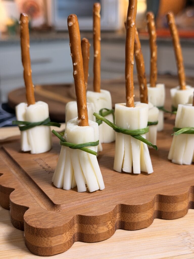 String cheese brooms with pretzel stick handles and green onion ties are arranged upright on a wooden board in a kitchen setting—one of 10 Easy Halloween Ideas perfect for your next spooky celebration.