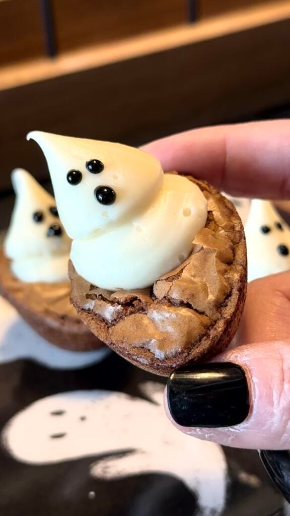 A hand holds a brownie bite topped with white frosting shaped like a ghost with black eyes, making it a perfect addition to your list of 10 Easy Halloween Ideas. Another similar treat is blurred in the background.