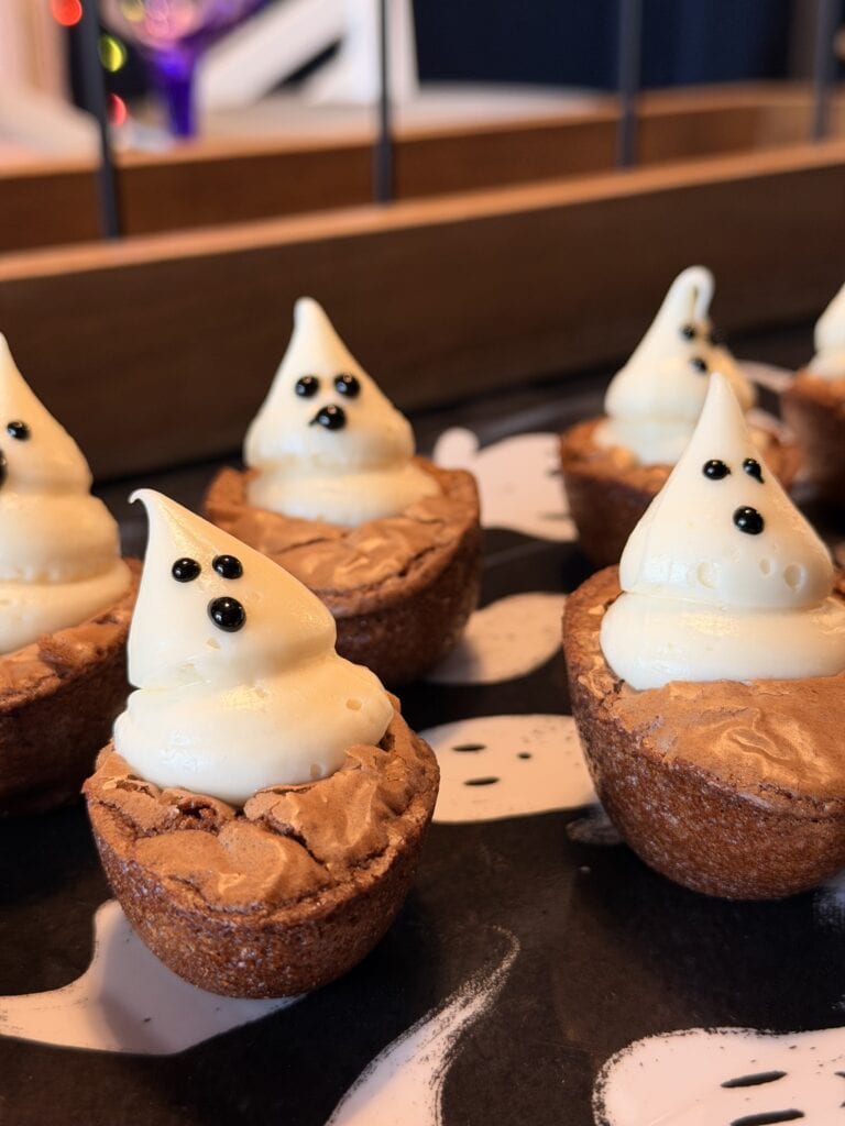 Mini brownie bites topped with white frosting shaped like ghosts, with small black eyes and mouths, arranged on a tray with a ghost pattern—these spooky treats are one of 10 easy Halloween ideas perfect for your next party.