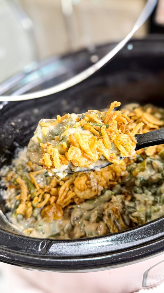 A close-up of a spoonful of green bean casserole topped with crispy fried onions, held over a slow cooker filled with more Monster Mix casserole.