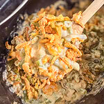 A wooden spoon lifts a serving of creamy green bean casserole topped with crispy fried onions and a hint of Monster Mix from a dark dish.