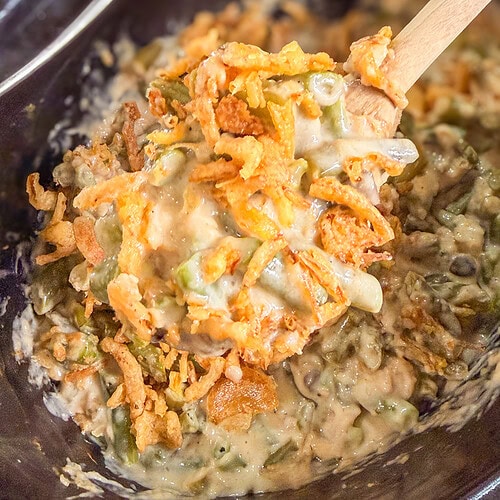 A wooden spoon lifts a serving of creamy green bean casserole topped with crispy fried onions and a hint of Monster Mix from a dark dish.