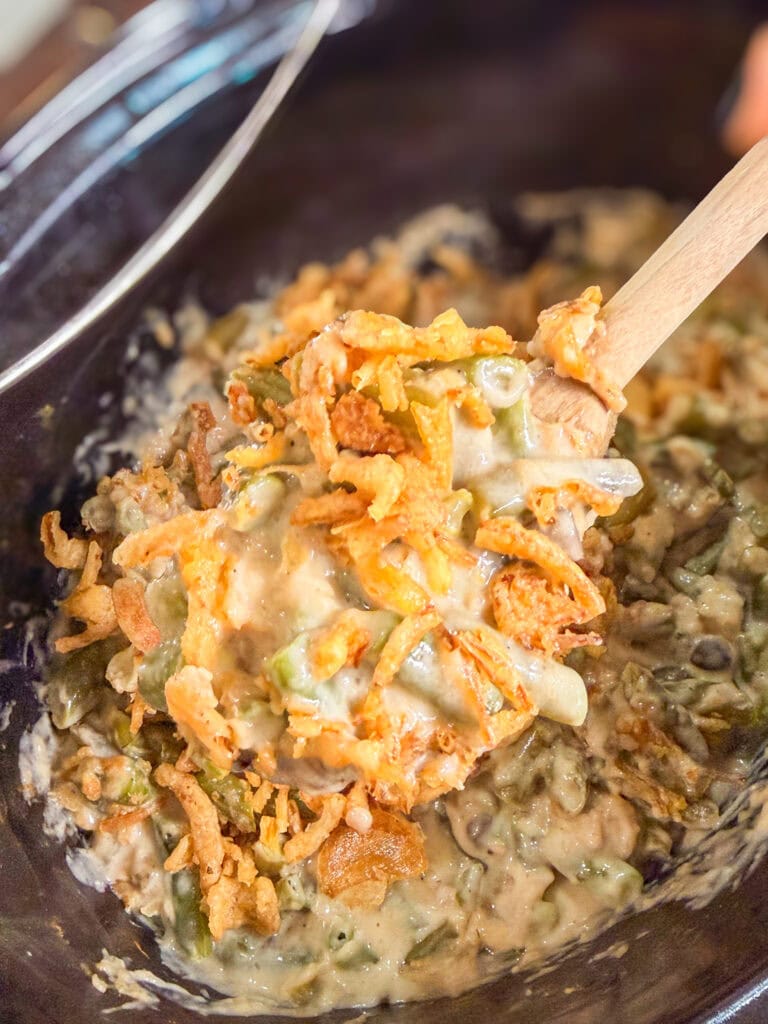 A wooden spoon lifts a serving of creamy green bean casserole topped with crispy fried onions and a hint of Monster Mix from a dark dish.
