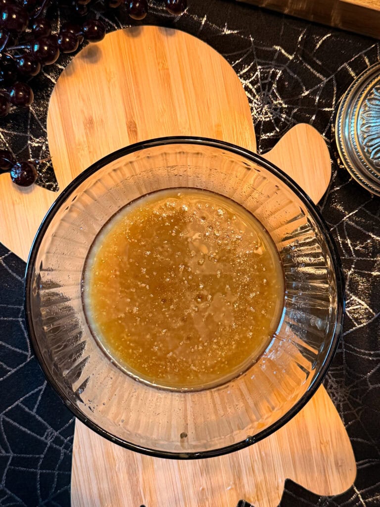 A glass bowl with a brown Monster Mix sits on a wooden cutting board shaped like a gingerbread person, with grapes and a spiderweb-patterned tablecloth nearby.