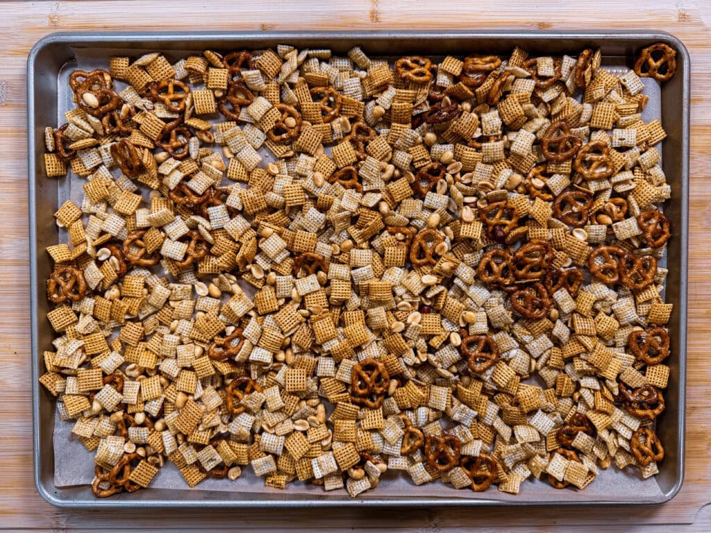 A baking sheet filled with homemade Monster Mix Chex mix, including cereal squares, mini pretzels, and peanuts—all toasted and seasoned—sits on a wooden surface.