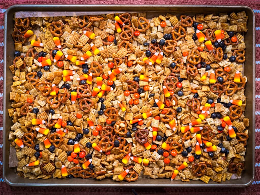 A baking sheet filled with Monster Mix, featuring a snack blend of Chex cereal, pretzels, candy corn, and chocolate candies, spread out evenly. The tray is set on a red and white striped cloth.