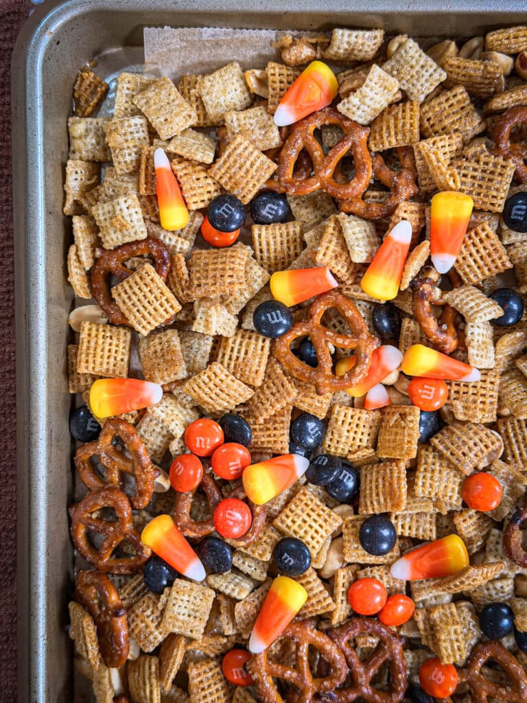 A baking tray filled with Chex cereal, pretzels, candy corn, peanuts, and colorful chocolate candies comes together to create a festive Monster Mix snack blend.
