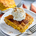 Squares of Pumpkin Cheesecake Bars on a white plate, topped with whipped cream and sprinkled with cinnamon.