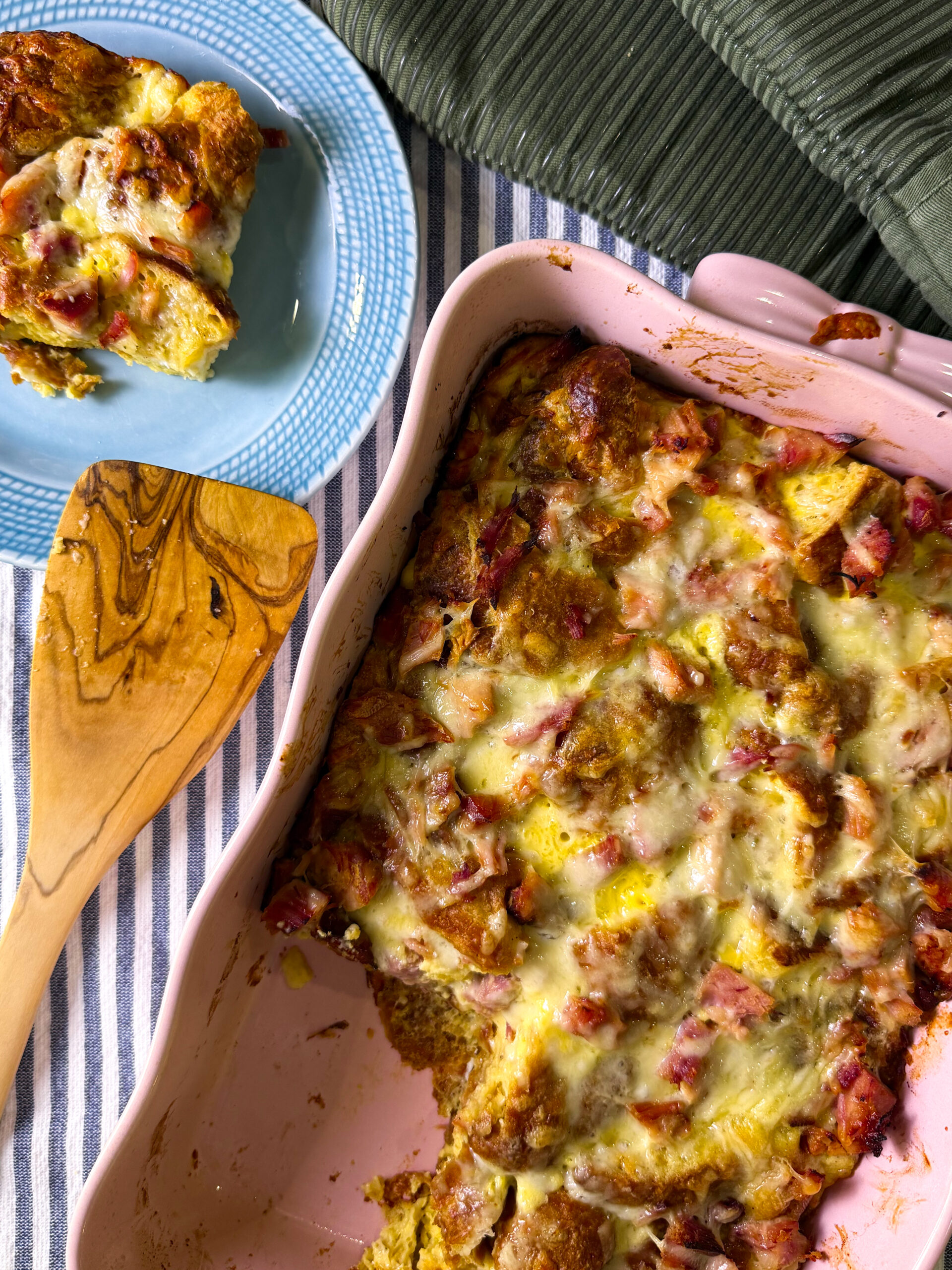 A baked casserole with melted cheese and diced ham in a pink dish, served beside roasted balsamic honey glazed carrots. A portion sits on a blue plate, all arranged on a striped cloth with a green napkin nearby.
