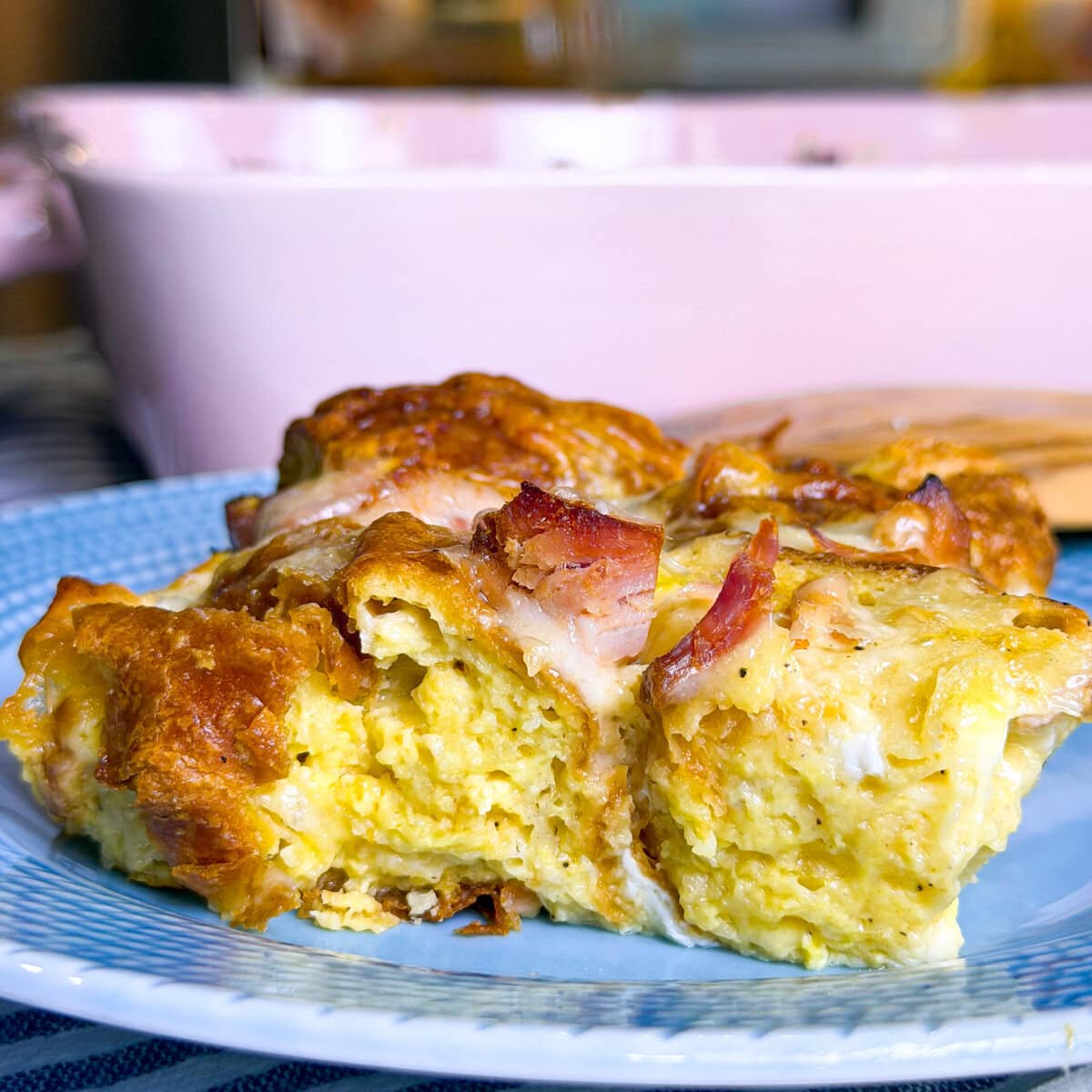 A close-up of a breakfast casserole slice on a blue plate, layered with egg, cheese, and ham, with a golden brown, slightly crispy top. Roasted Balsamic Honey Glazed Carrots add a colorful touch beside the casserole. A baking dish is blurred in the background.
