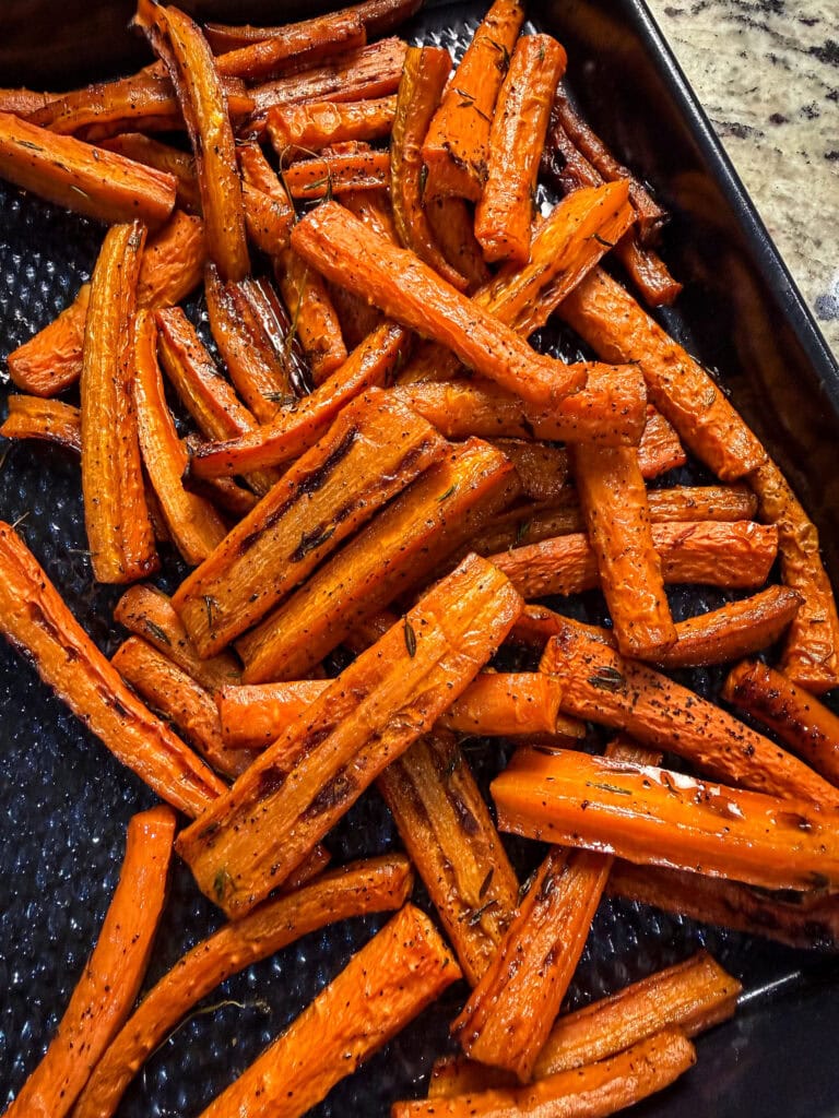 Roasted Balsamic Honey Glazed Carrots, seasoned with herbs and spices, are spread out on a dark, textured baking tray. The carrots appear golden-brown and slightly charred for a delicious finish.
