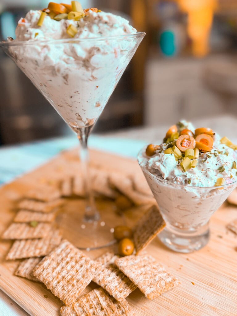 Dirty martini inspired dip made with cream cheese, sour cream, green olives, parsley, seasonings, and blue cheese served in a short martini glass with wheat crackers on the side