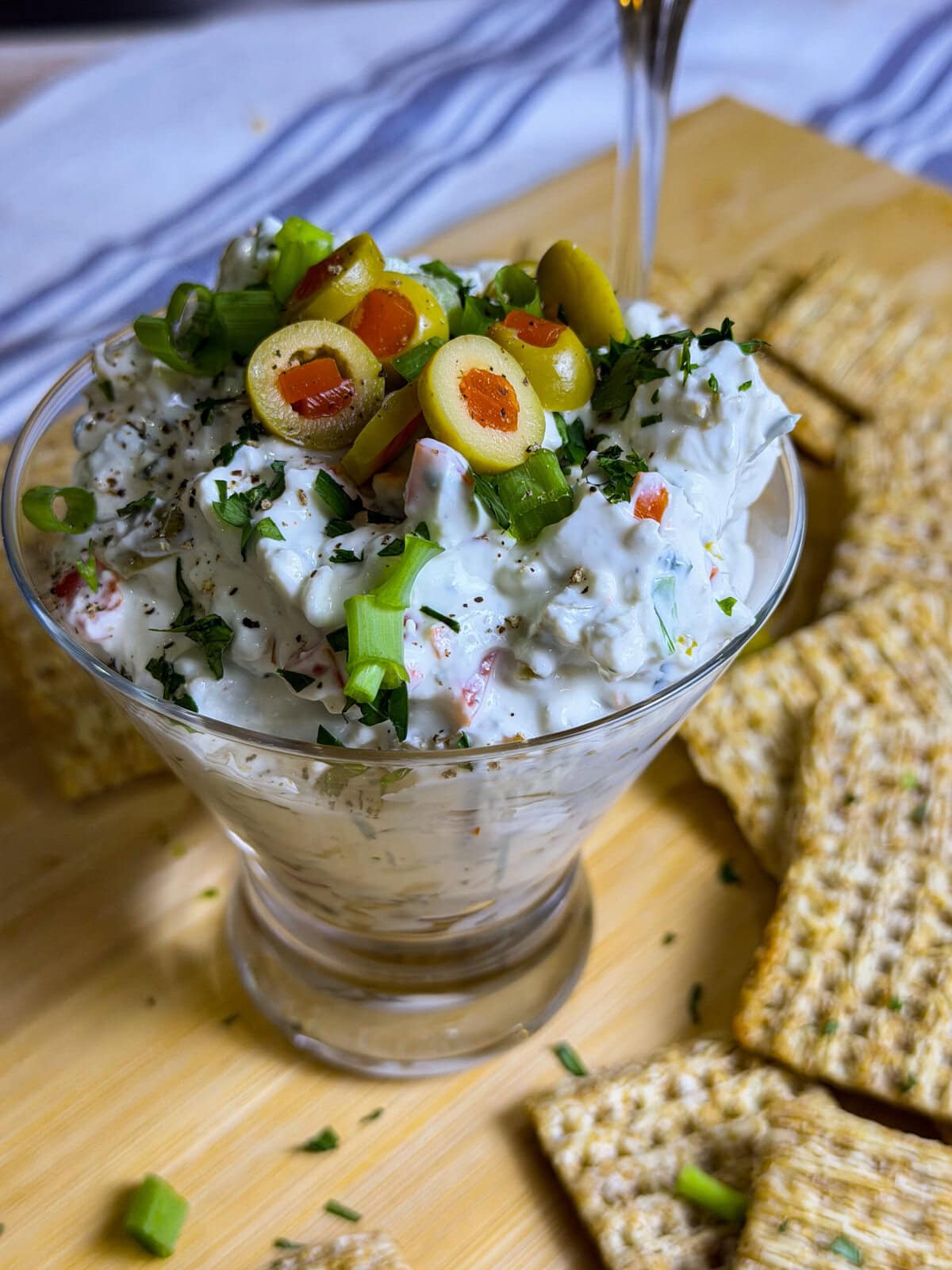 Dirty martini inspired dip made with cream cheese, sour cream, green olives, parsley, seasonings, and blue cheese served in a short martini glass with wheat crackers on the side