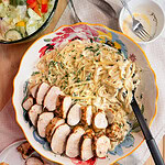 A large floral bowl with creamy fettuccine tossed in Copycat Olive Garden Alfredo Sauce is topped with sliced grilled chicken breast, served alongside salad, extra chicken slices, garlic bread, and a small bowl of dressing.