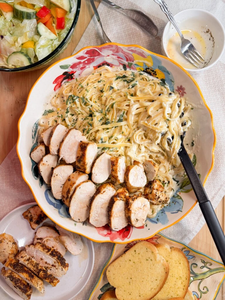 A large floral bowl with creamy fettuccine tossed in Copycat Olive Garden Alfredo Sauce is topped with sliced grilled chicken breast, served alongside salad, extra chicken slices, garlic bread, and a small bowl of dressing.