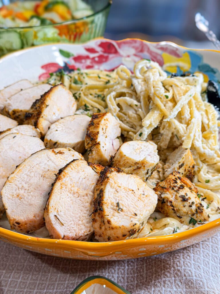 A colorful bowl filled with fettuccine pasta tossed in a creamy Copycat Olive Garden Alfredo Sauce, topped with several thick slices of seasoned, grilled chicken breast. A salad is partially visible in the background.