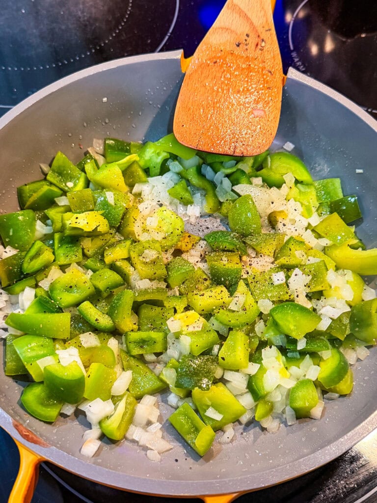 Chopped green bell peppers and diced onions are being sautéed in a gray pan on a stovetop, with a wooden spatula resting on the edge. There is visible black pepper sprinkled on the vegetables—perfectly preparing the flavorful base for a delicious Stuffed Pepper Casserole.