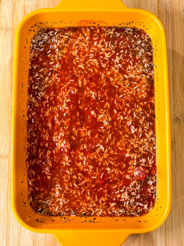 Yellow baking dish filled with an uncooked Stuffed Pepper Casserole rice and tomato sauce mixture, placed on a wooden surface.