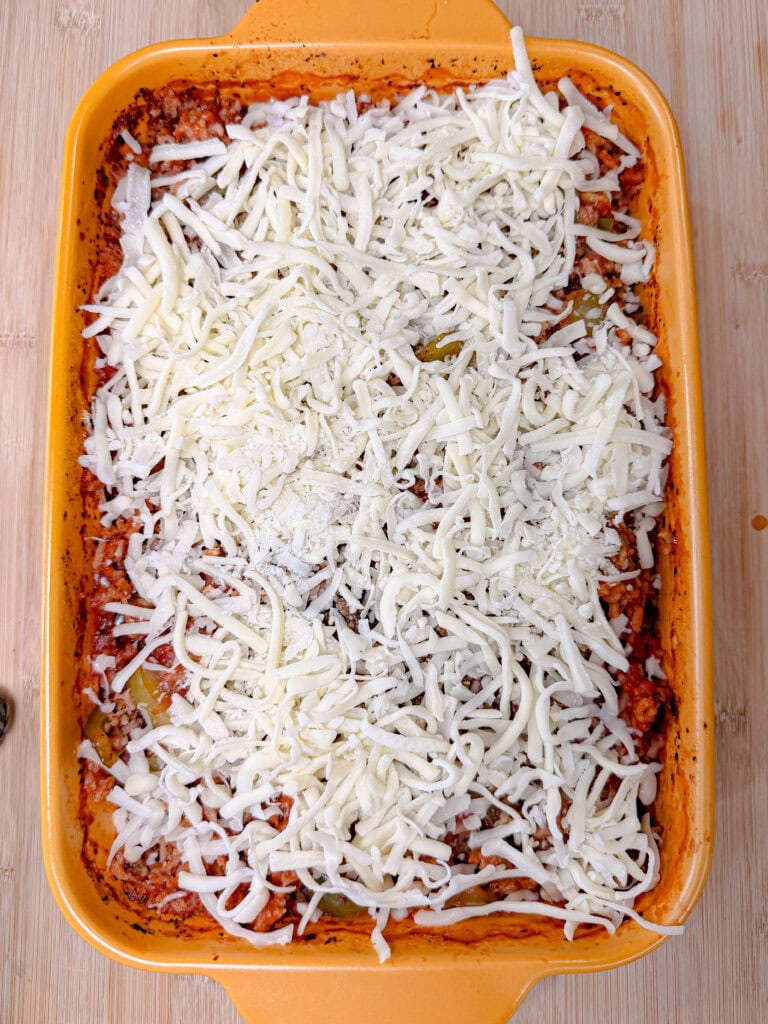 Rectangular orange baking dish filled with a delicious Stuffed Pepper Casserole, topped with a layer of shredded white cheese, ready to be baked, sitting on a light wooden surface.
