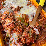 A yellow baking dish filled with Stuffed Pepper Casserole, featuring a cheesy blend of ground meat, rice, tomato sauce, and green bell peppers. A wooden spoon rests inside, with some of the hearty casserole already scooped out.