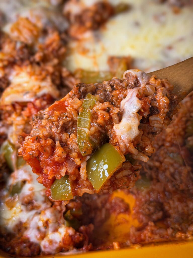 A wooden spoon holds a scoop of cheesy Stuffed Pepper Casserole with ground beef, rice, diced tomatoes, green bell peppers, and melted cheese, with more casserole visible in the background.