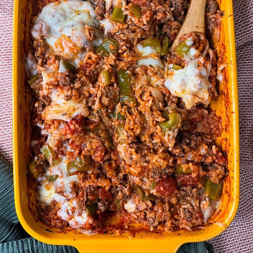 A yellow baking dish filled with a cheesy Stuffed Pepper Casserole made of rice, ground beef, green bell peppers, tomato sauce, and melted cheese, with a wooden spoon scooping some out.