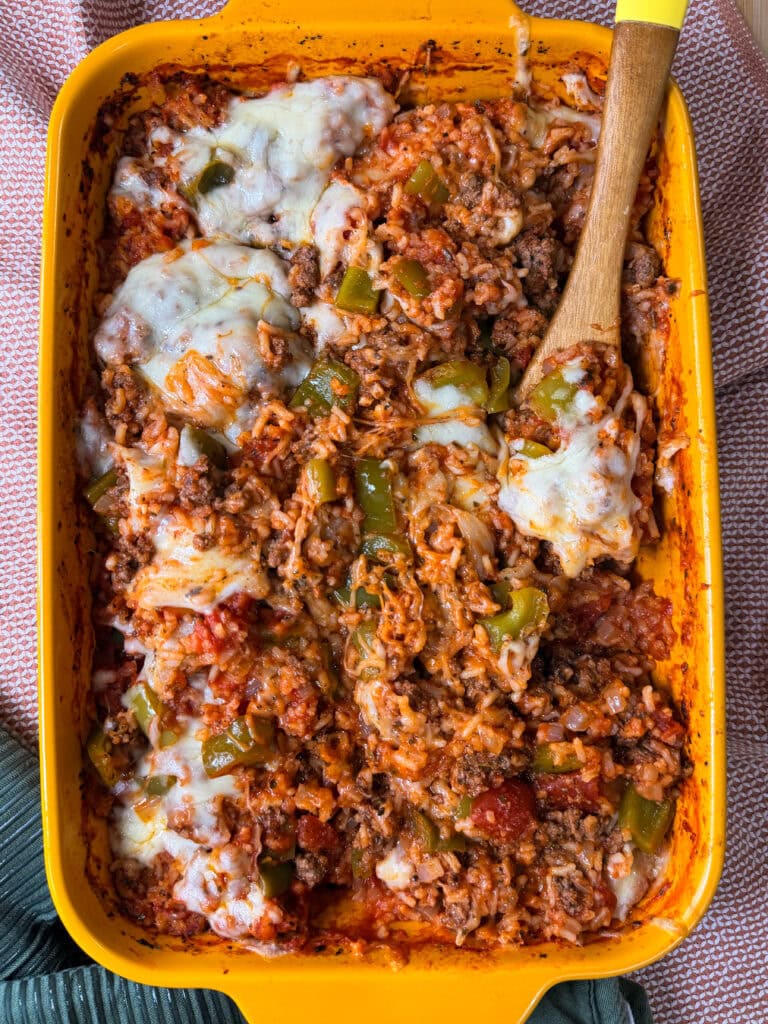 A yellow baking dish filled with a cheesy Stuffed Pepper Casserole made of rice, ground beef, green bell peppers, tomato sauce, and melted cheese, with a wooden spoon scooping some out.
