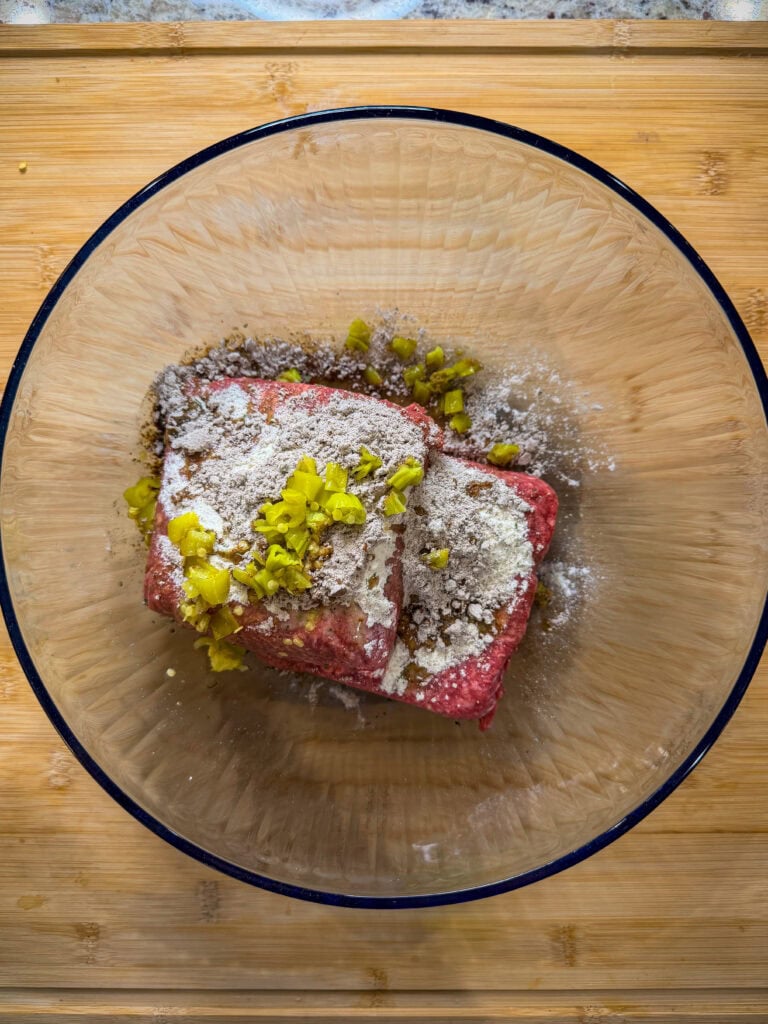 A glass bowl filled with ground meat, diced green peppers, and various powdered seasonings sits on a wooden cutting board—perfect for preparing Sheet Pan Mississippi Burger Sliders.