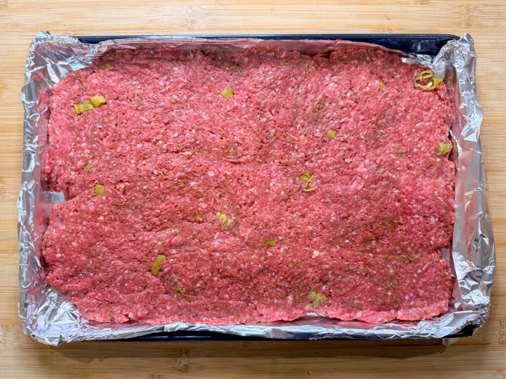 Raw ground beef is evenly spread out in a rectangular layer on a foil-lined baking sheet, with small pieces of green peppers mixed in—perfect for making Sheet Pan Mississippi Burger Sliders. The baking sheet rests on a wooden surface.