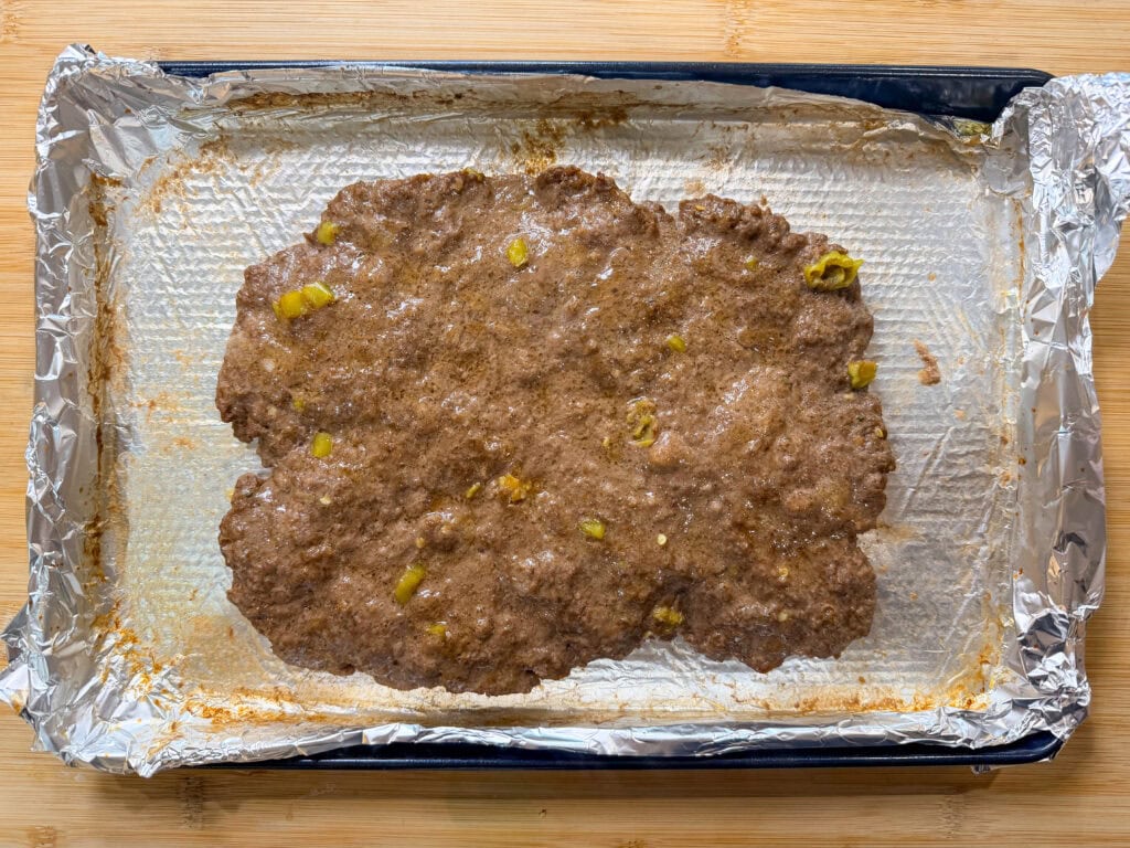 A sheet pan lined with aluminum foil holds a spread-out mixture of uncooked ground meat and chopped green peppers, ready to become delicious Sheet Pan Mississippi Burger Sliders. The pan sits on a wooden surface.