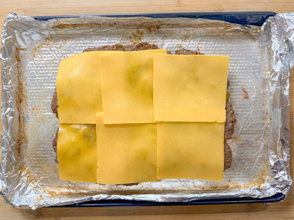 A sheet pan lined with foil holds a rectangular layer of cooked ground beef topped with six overlapping slices of yellow cheese, perfect for making Sheet Pan Mississippi Burger Sliders.