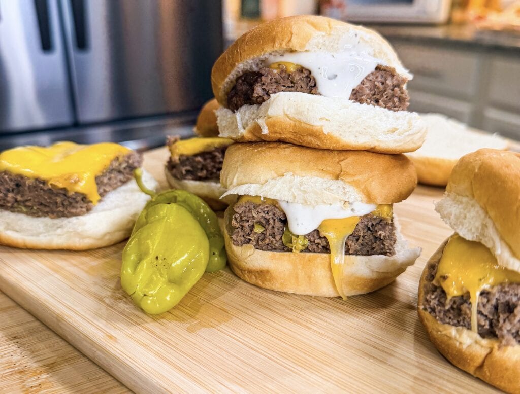 Three cheeseburgers with beef patties, cheddar cheese, and white sauce are stacked on a wooden board beside sliced pickles, capturing the tasty appeal of Sheet Pan Mississippi Burger Sliders in a cozy kitchen setting.