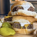 Two freshly made Sheet Pan Mississippi Burger Sliders with melted cheese and white sauce are stacked on a wooden board next to green peppers, nestled in soft white buns.