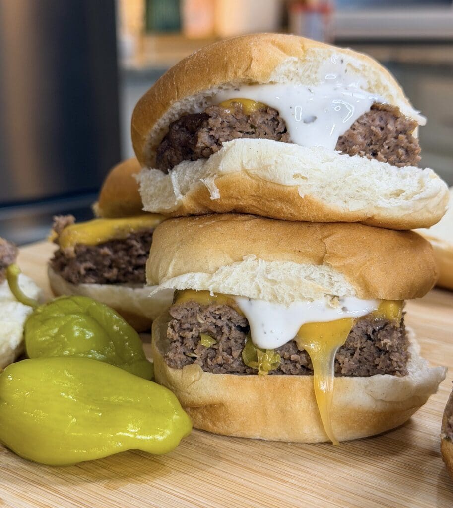 Two freshly made Sheet Pan Mississippi Burger Sliders with melted cheese and white sauce are stacked on a wooden board next to green peppers, nestled in soft white buns.