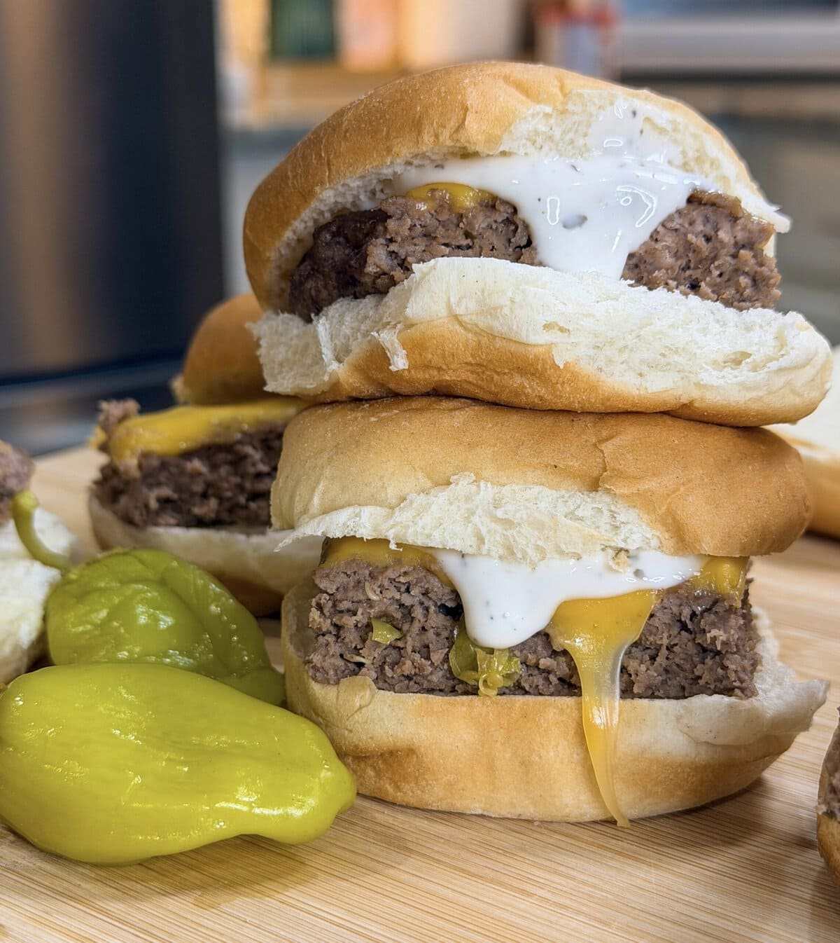 Two freshly made Sheet Pan Mississippi Burger Sliders with melted cheese and white sauce are stacked on a wooden board next to green peppers, nestled in soft white buns.