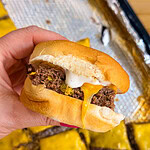 A hand holds a Sheet Pan Mississippi Burger Slider with a beef patty, melted cheese, pickles, and sauce in a soft bun. In the background, more sliders rest on a foil-lined tray.