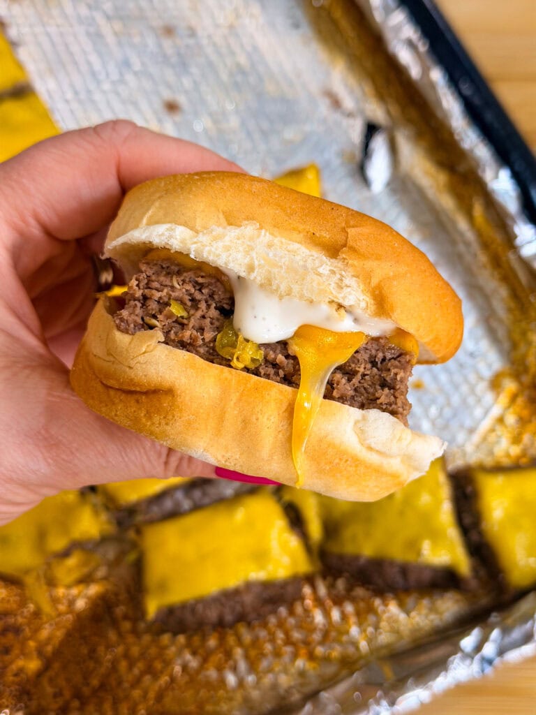A hand holds a Sheet Pan Mississippi Burger Slider with a beef patty, melted cheese, pickles, and sauce in a soft bun. In the background, more sliders rest on a foil-lined tray.
