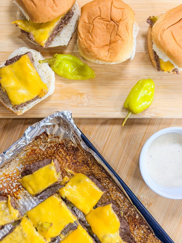 Sheet Pan Mississippi Burger Sliders with melted cheddar on a cutting board, served with pepperoncini peppers. A foil-lined baking tray with more sliders and a cup of white dipping sauce rest on a wooden surface.