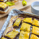 Sheet Pan Mississippi Burger Sliders with melted cheddar on beef patties are arranged on a tray and wooden board, accompanied by yellow peppers and a small dish of white dipping sauce.
