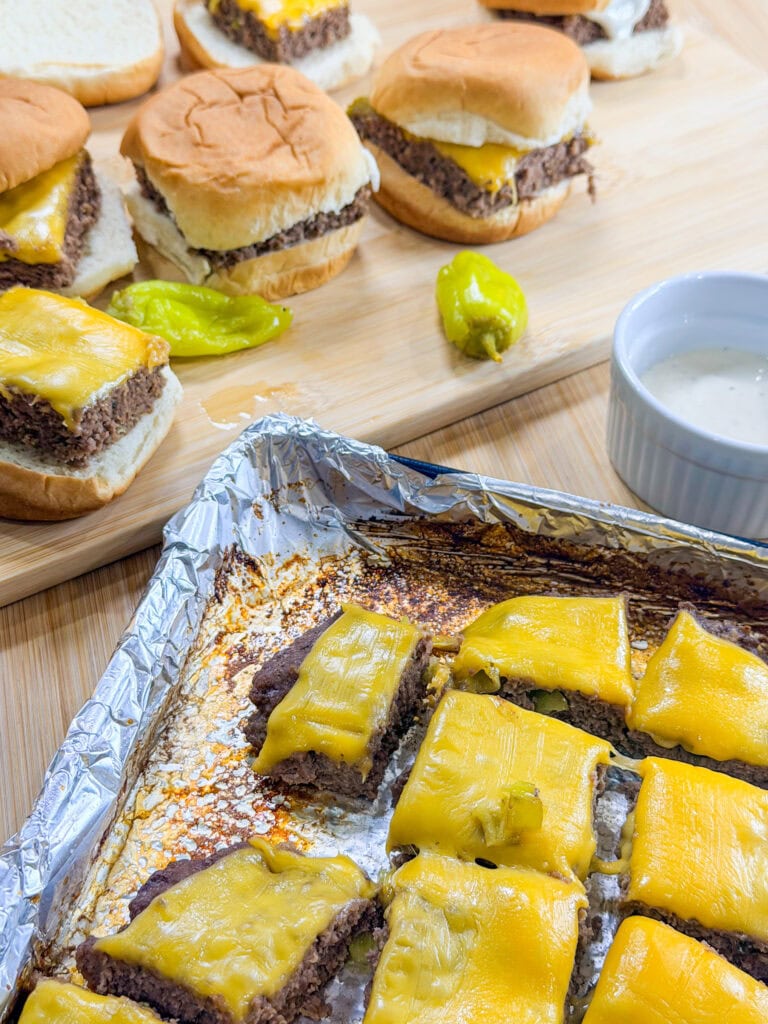 Sheet Pan Mississippi Burger Sliders with melted cheddar on beef patties are arranged on a tray and wooden board, accompanied by yellow peppers and a small dish of white dipping sauce.