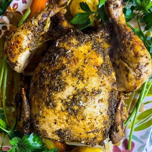 A Mardi Gras Crock Pot Chicken, whole roasted and seasoned with herbs, is garnished with fresh parsley and lemon wedges, then served on a floral-patterned plate with carrots and celery.