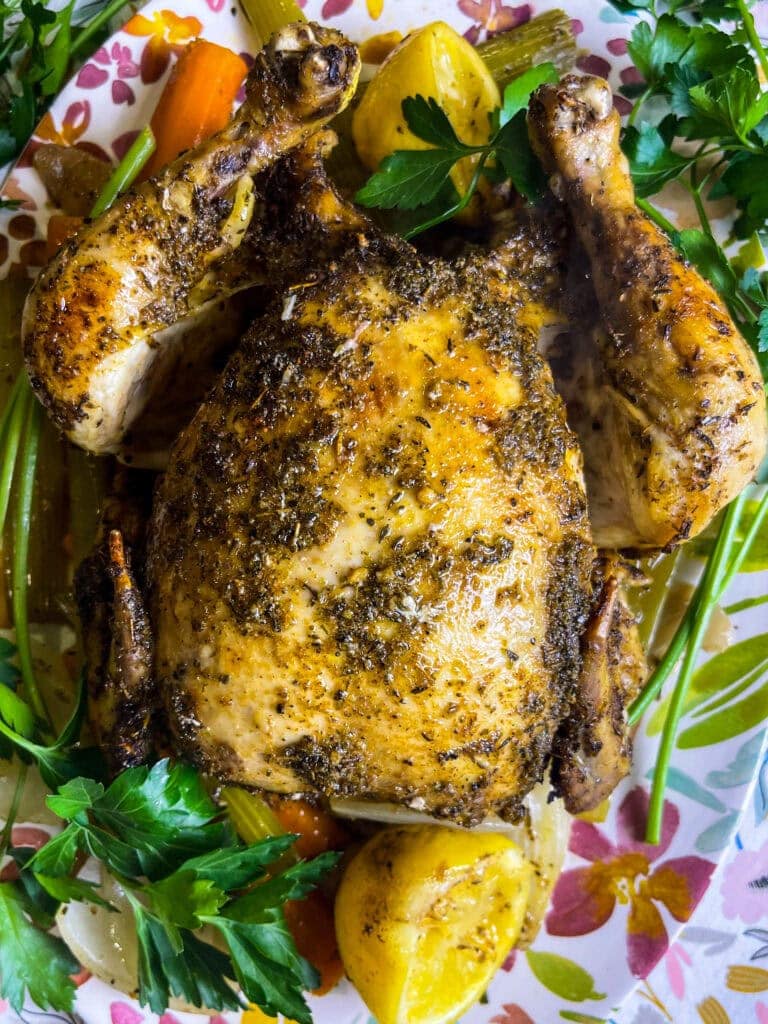 A Mardi Gras Crock Pot Chicken, whole roasted and seasoned with herbs, is garnished with fresh parsley and lemon wedges, then served on a floral-patterned plate with carrots and celery.