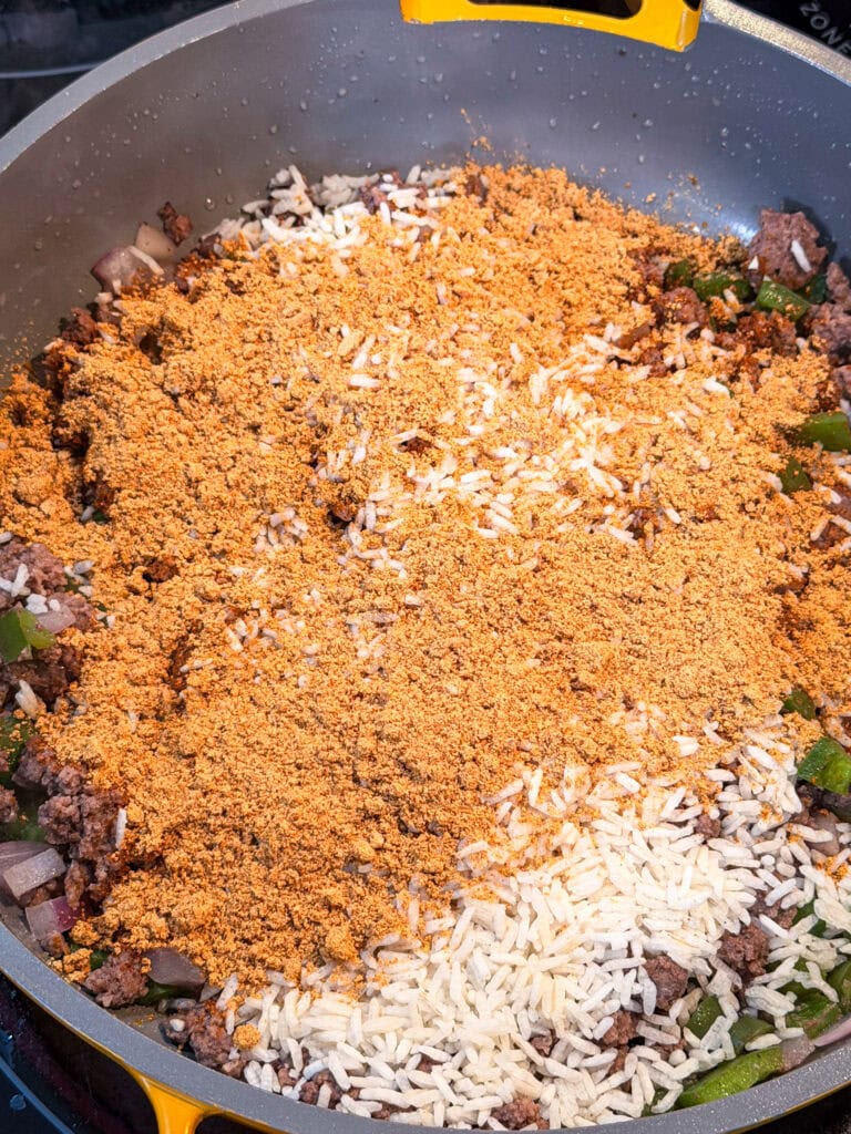 A Taco Skillet filled with browned ground beef, chopped onions, green peppers, white rice, and a generous layer of taco seasoning sits ready to be mixed together.