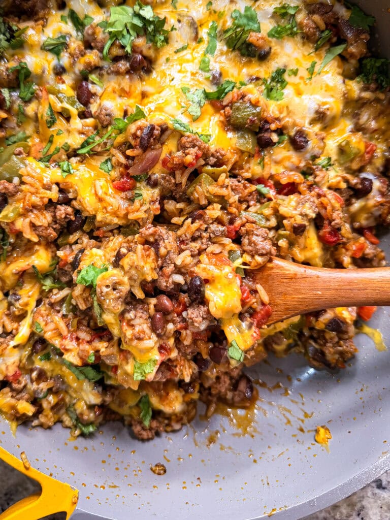 A close-up of a Taco Skillet filled with cheesy ground beef, rice, black beans, tomatoes, and herbs, being stirred with a wooden spoon.