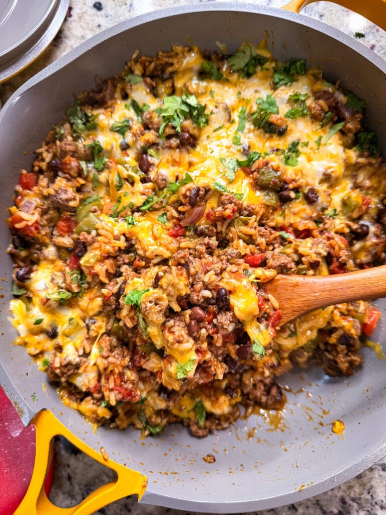 A Taco Skillet filled with a cheesy beef, rice, black bean, and vegetable mixture, topped with melted cheese and fresh cilantro, with a wooden spoon mixing the contents.