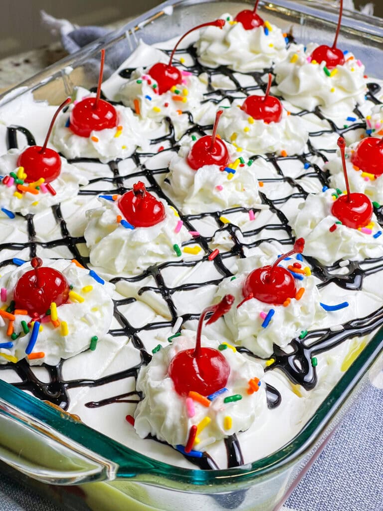 poke cake made with banana cake mix, strawberry jam, vanilla pudding mixed with crushed pineapples and topped with whipped cream and chocolate sauce